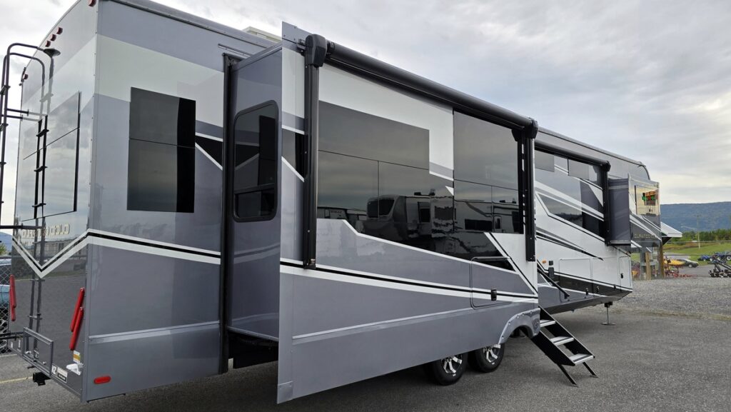 Modern gray fifth-wheel RV with extended slide-outs and stairs parked on a paved lot under a cloudy sky.