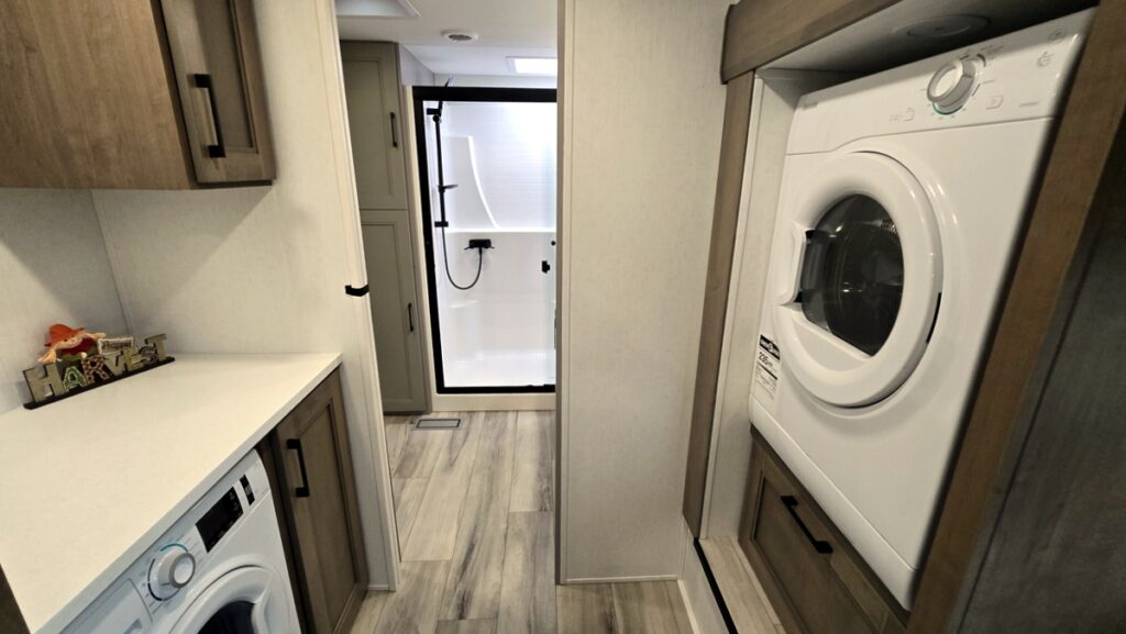 Compact laundry area inside a modern RV featuring a stacked washer and dryer next to a small hallway leading to a shower room.